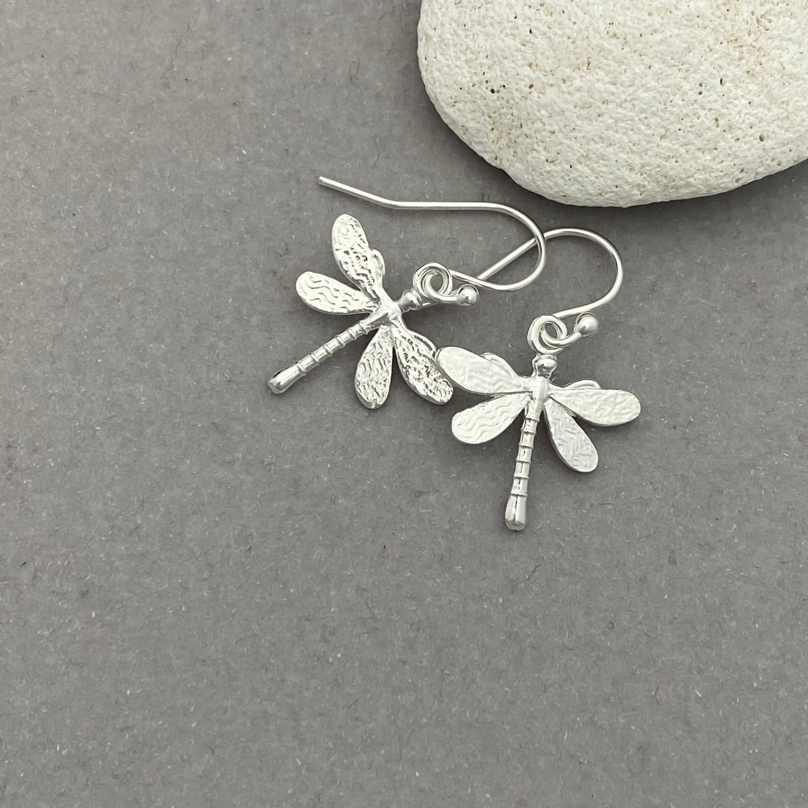 Silver dragonfly earrings on a gray surface with a stone in the background
