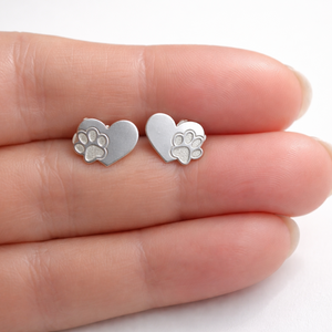 Silver heart-shaped earrings with paw prints held between fingers on a white background