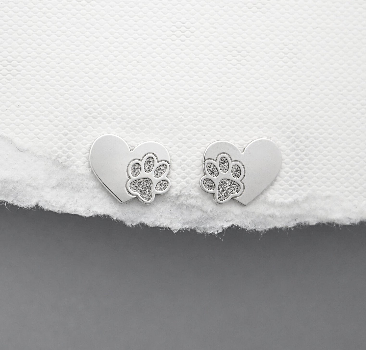 Heart-shaped silver earrings with paw prints on a white fabric background